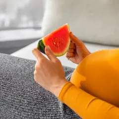 Ciężarna kobieta w pomarańczowej bluzce trzyma kawałek soczystego arbuza.
