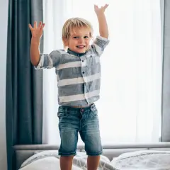 Radosny chłopiec w dżinsach i pasiastym T-shircie skacze na łóżku, ćwicząc swoją motorykę.
