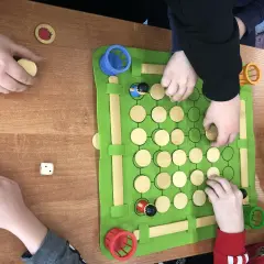 Dzieci grają w grę planszową, która jest częścią zajęć korekcyjno kompensacyjnych. Na zielonej macie znajdują się drewniane pionki i kostka.