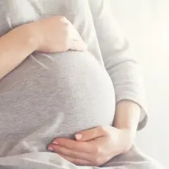 Brzuszek w 6 miesiącu ciąży, otulony szarym materiałem. Dłonie delikatnie go obejmują, tworząc ciepły krąg.