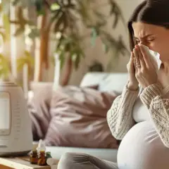 Ciężarna kobieta z chusteczką przy nosie szuka informacji: co na ból gardła w ciąży. Obok nawilżacz powietrza i buteleczki z lekami.