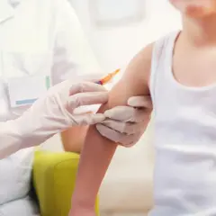Dziecko otrzymuje szczepionkę 6w1. Pielęgniarka w białym fartuchu i rękawiczkach podaje zastrzyk w ramię malucha.