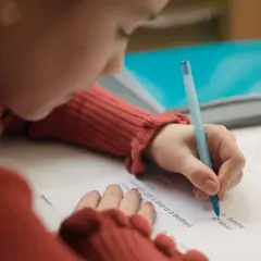 Dziecko w czerwonym swetrze pisze długopisem na kartce. Czy to zadanie z języka angielskiego, od jakiej średniej jest 3?