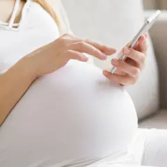 Kobieta w 34. tygodniu ciąży przegląda coś na telefonie, siedząc wygodnie na kanapie.