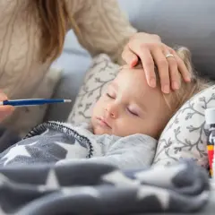 Mama sprawdza temperaturę chorego dziecka. Na stoliku leki, a maluch śpi pod gwiazdkową kołderką. L4 na dziecko.