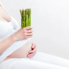 Ciężarna kobieta w białej sukience trzyma pęczek zielonych szparagów, symbol zdrowej diety w ciąży.