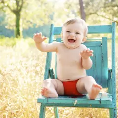 Uśmiechnięte dziecko w czerwonych majteczkach, kiedy dziecko siada na turkusowym krześle w słonecznym ogrodzie.