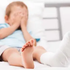 Chłopiec z zabandażowaną nogą, ukrywa twarz w dłoniach. Może to być skutek wypadku lub objaw krzywicy.