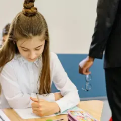 Dziewczynka skupiona na zadaniu podczas testu trzecioklasisty. Nauczyciel stoi obok, trzymając okulary.