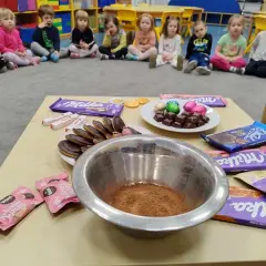 Dzieci w przedszkolu świętują dzień czekolady. Na stole czekolady Milka, Kinder, ciastka i miska z kakao.