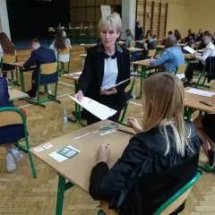 Napięcie w sali gimnastycznej, kiedy rozdawane są arkusze. Uczniowie czekają na wyniki egzaminu ósmoklasisty, a nauczycielka czuwa nad przebiegiem.