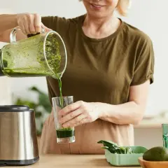 Kobieta nalewa zielony koktajl położnej do szklanki. Obok stoi blender i miska z owocami.