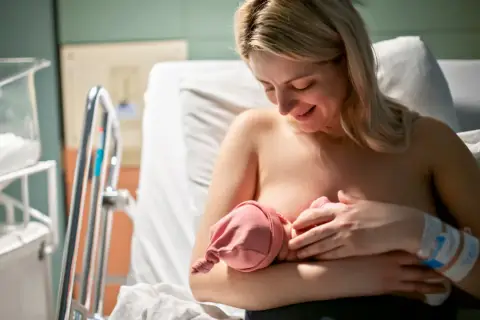 Uśmiechnięta mama karmi piersią noworodka w szpitalu. Choć nie widać tego bezpośrednio, może to być moment, gdy pojawia się grzybica sutka, wymagająca uwagi.