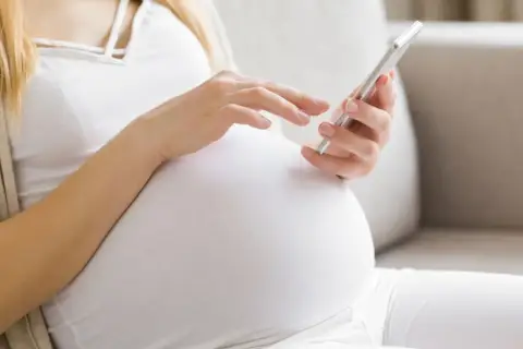 Kobieta w 34. tygodniu ciąży przegląda coś na telefonie, siedząc wygodnie na kanapie.