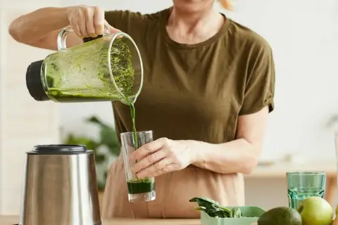 Kobieta nalewa zielony koktajl położnej do szklanki. Obok stoi blender i miska z owocami.