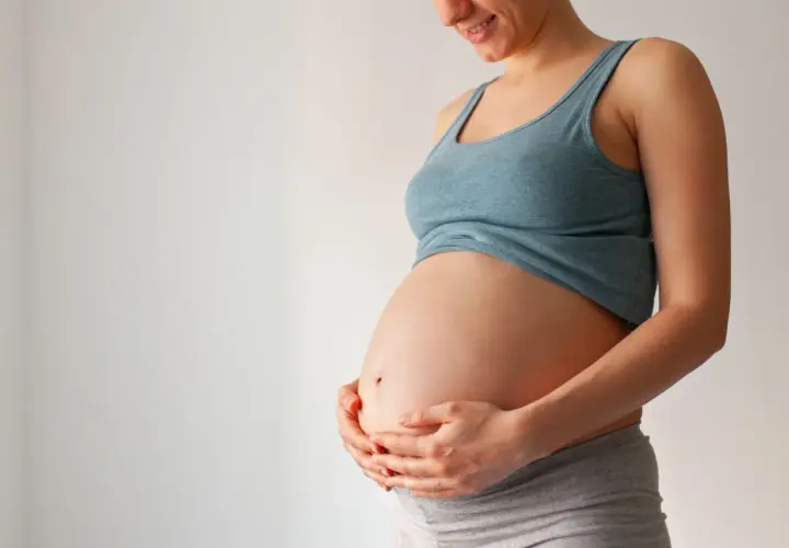 Uśmiechnięta ciężarna kobieta trzyma dłonie na brzuchu. Wkrótce może pojawić się czop śluzowy.