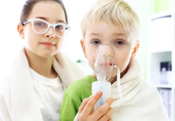 Dziecko z maską inhalacyjną, wspierane przez siostrę w okularach. Dzieci przechodzą inhalacje z soli fizjologicznej.