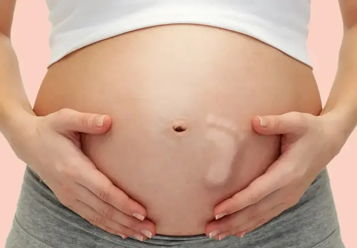 Brzuszek w 30 tygodniu ciąży, z widocznym odciskiem stópki dziecka. Ręce mamy delikatnie obejmują rosnący brzuch.