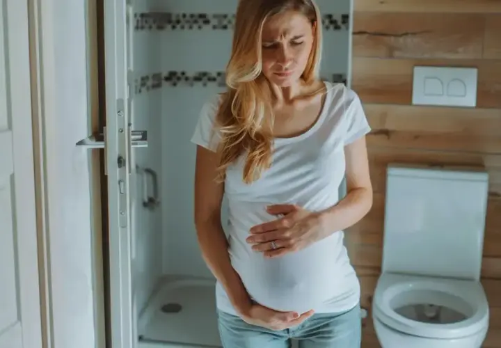 Kobieta w ciąży z wyrazem bólu na twarzy trzyma się za brzuch. W tle łazienka z toaletą. Ból podbrzusza w ciąży.