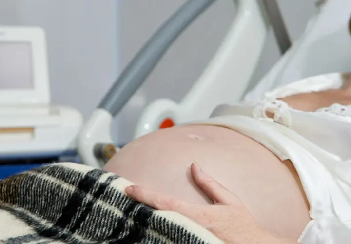 Kobieta w szpitalnym łóżku, z ręką na brzuchu, czeka na poród. Obok monitor KTG, który pomaga ocenić stan dziecka i może sugerować, jak przyspieszyć poród.