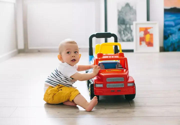 Uśmiechnięte niemowlę w żółtych spodenkach bawi się czerwono-żółtą ciężarówką na drewnianej podłodze.