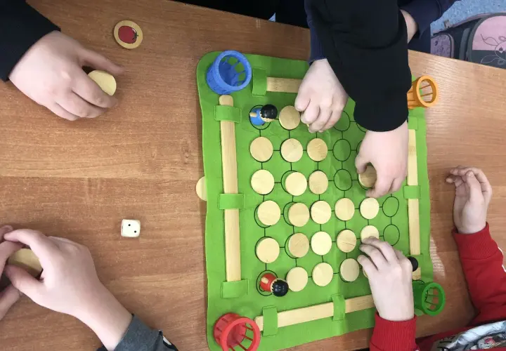 Dzieci grają w grę planszową, która jest częścią zajęć korekcyjno kompensacyjnych. Na zielonej macie znajdują się drewniane pionki i kostka.