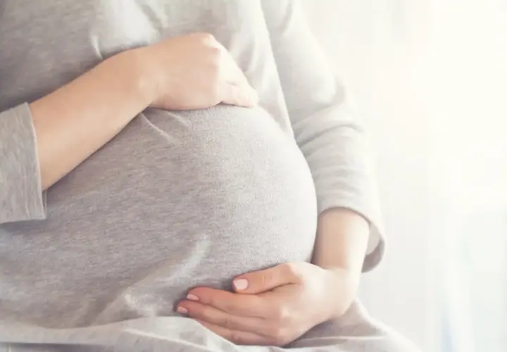 Brzuszek w 6 miesiącu ciąży, otulony szarym materiałem. Dłonie delikatnie go obejmują, tworząc ciepły krąg.