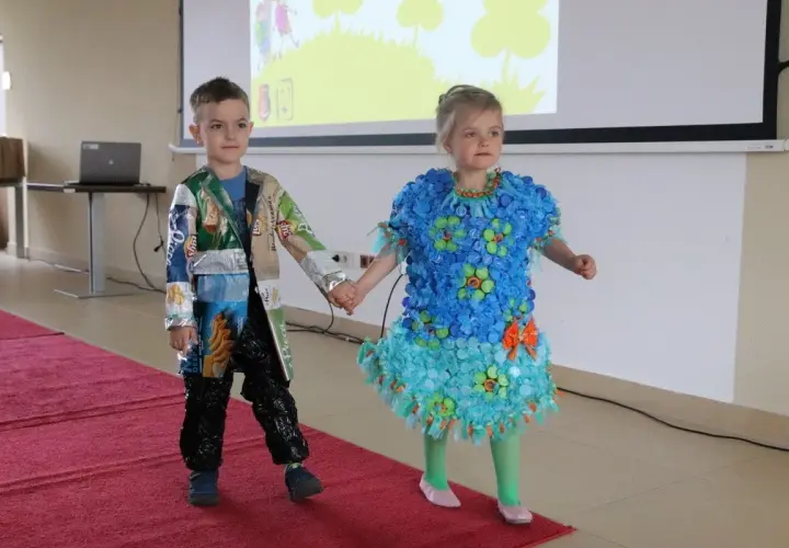 Dzieci prezentują stroje ekologiczne. Dziewczynka w niebieskiej sukience z nakrętek, chłopiec w kurtce z opakowań. Inspiracja, jak zrobić strój ekologiczny dla dziewczynki.