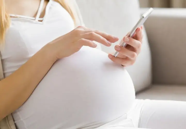 Kobieta w 34. tygodniu ciąży przegląda coś na telefonie, siedząc wygodnie na kanapie.