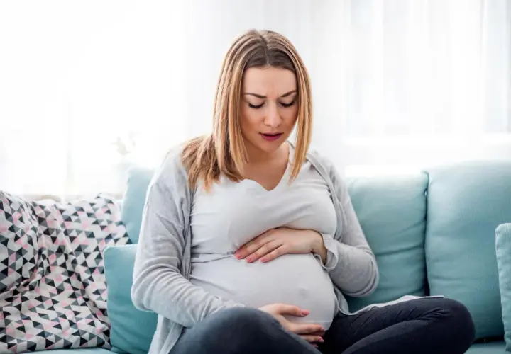Kobieta w ciąży siedzi na kanapie, trzymając się za brzuch. Jej twarz wyraża dyskomfort, być może to skurcze przepowiadające.