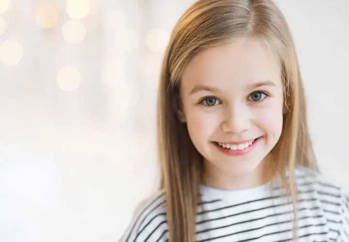 Uśmiechnięta dziewczynka o blond włosach, jakby inspirowana nowoczesnymi imionami dla dziewczyn, z rozmazanym tłem pełnym świateł.