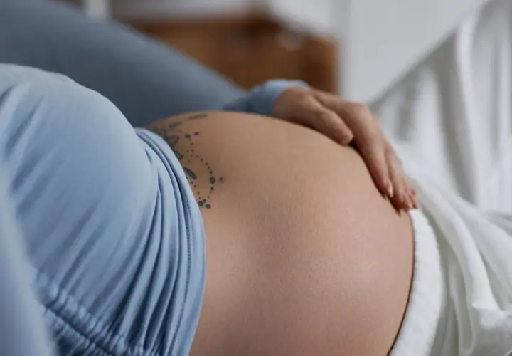 Dłoń delikatnie spoczywa na **twardym brzuchu w ciąży**, ozdobionym subtelnym tatuażem.