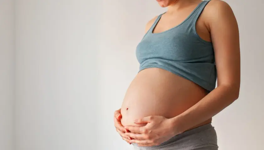 Uśmiechnięta ciężarna kobieta trzyma dłonie na brzuchu. Wkrótce może pojawić się czop śluzowy.