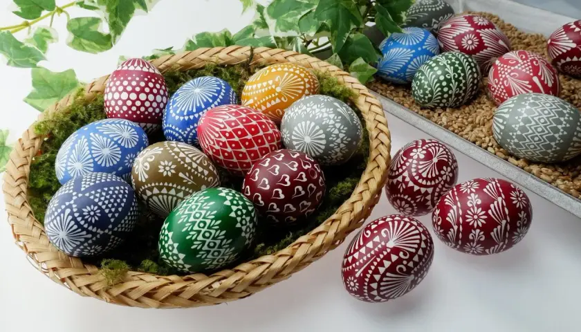 Kosz pełen barwnych pisanki kraszanki z tradycyjnymi wzorami. Wśród nich jajka zdobione białymi ornamentami: kwiatami, serduszkami i geometrycznymi motywami.