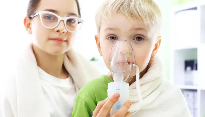 Dziecko z maską inhalacyjną, wspierane przez siostrę w okularach. Dzieci przechodzą inhalacje z soli fizjologicznej.