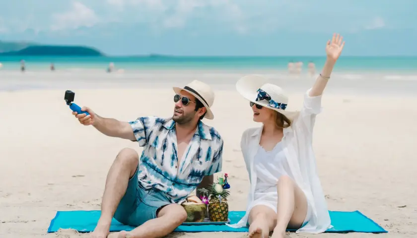 Para w letnich strojach robi sobie selfie na plaży, z napojami i owocami. To idealne wczasy pod gruszą!