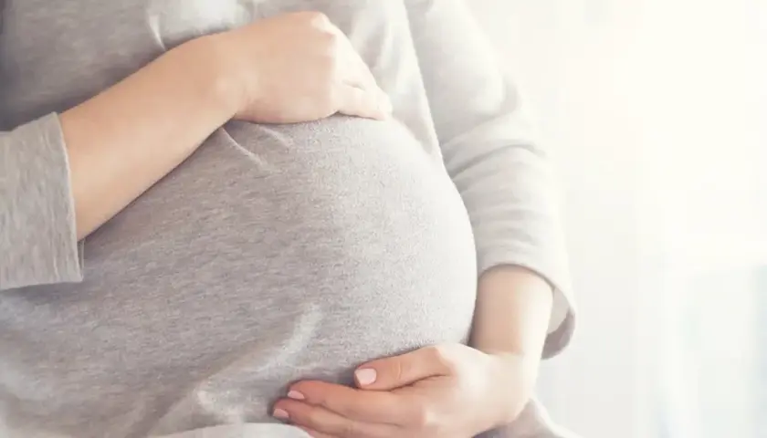 Brzuszek w 6 miesiącu ciąży, otulony szarym materiałem. Dłonie delikatnie go obejmują, tworząc ciepły krąg.