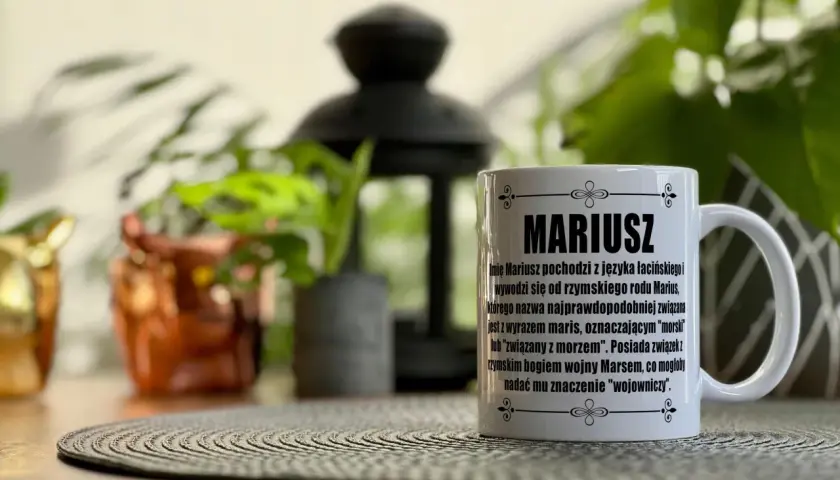 Biały kubek z napisem "MARIUSZ" i opisem pochodzenia imienia. W tle rośliny i ozdobna lampa.