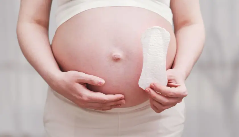 Ciąża podczas okresu? Kobieta trzyma podpaskę na tle swojego ciążowego brzucha, sugerując pytanie o płodność.