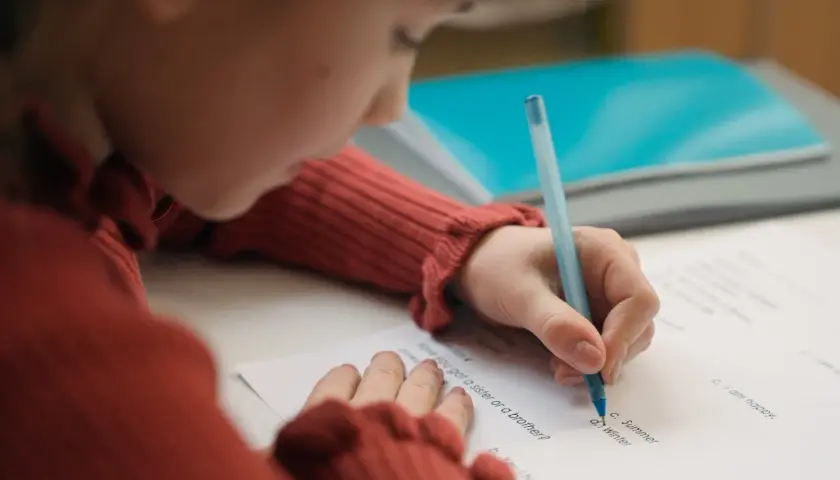 Dziecko w czerwonym swetrze pisze długopisem na kartce. Czy to zadanie z języka angielskiego, od jakiej średniej jest 3?