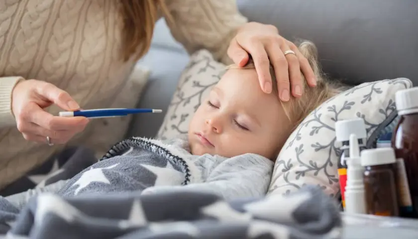 Mama sprawdza temperaturę chorego dziecka. Na stoliku leki, a maluch śpi pod gwiazdkową kołderką. L4 na dziecko.