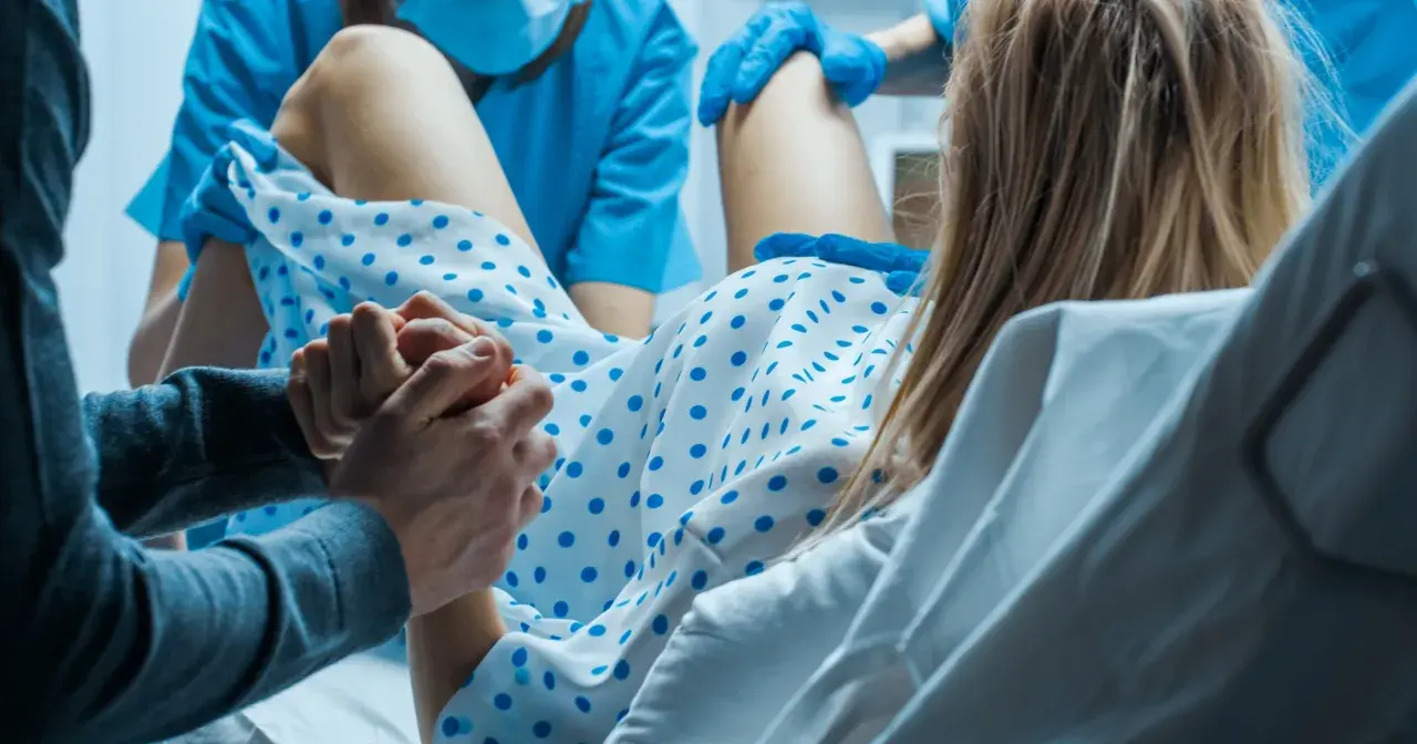 Wsparcie bliskiej osoby podczas porodu. Kobieta trzyma dłoń partnera, otoczona personelem medycznym. To nie jest balonik do porodu, lecz chwila narodzin.