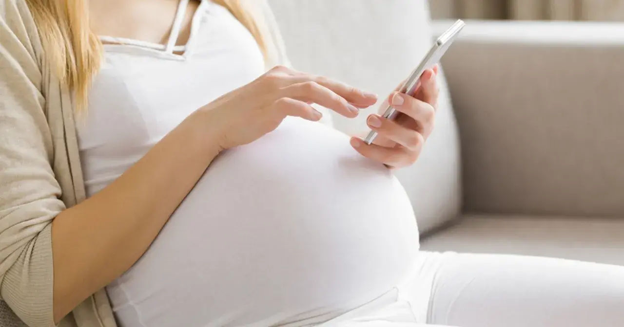 Kobieta w 34. tygodniu ciąży przegląda coś na telefonie, siedząc wygodnie na kanapie.