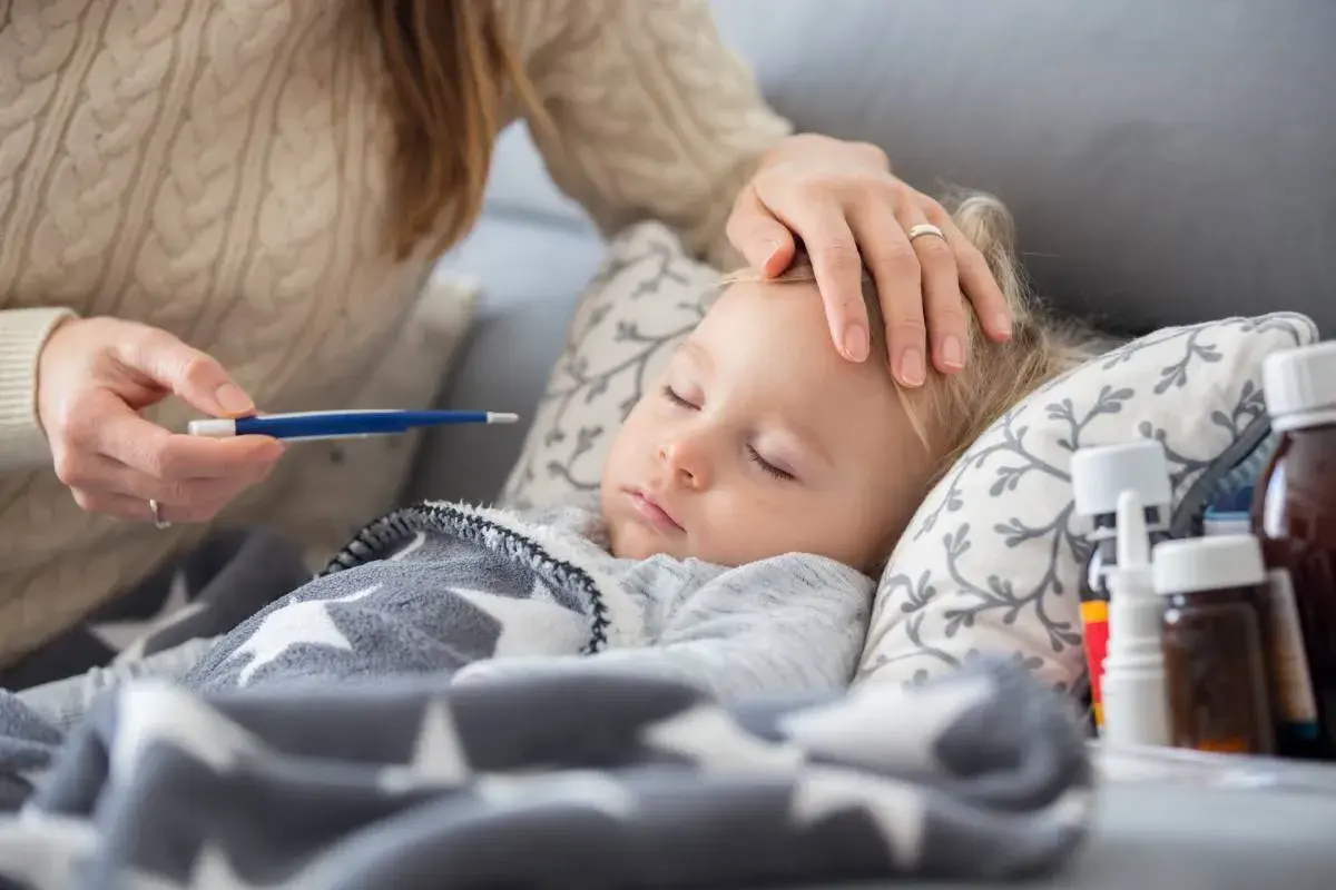 Mama sprawdza temperaturę chorego dziecka. Na stoliku leki, a maluch śpi pod gwiazdkową kołderką. L4 na dziecko.