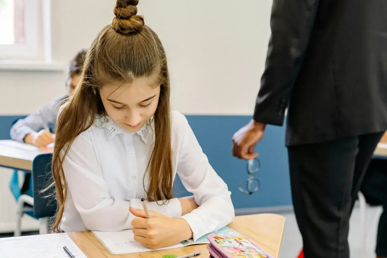 Dziewczynka skupiona na zadaniu podczas testu trzecioklasisty. Nauczyciel stoi obok, trzymając okulary.