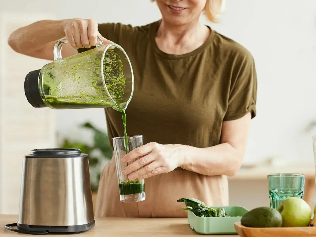 Kobieta nalewa zielony koktajl położnej do szklanki. Obok stoi blender i miska z owocami.