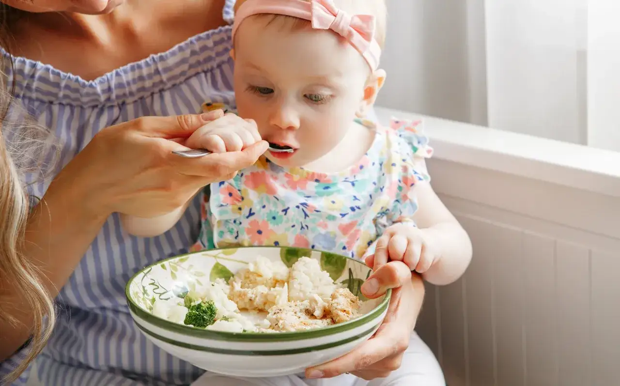 Mama karmi dziecko łyżeczką. Rozszerzanie diety to ważny etap, maluch próbuje nowych smaków: ryżu, brokułów i kurczaka.