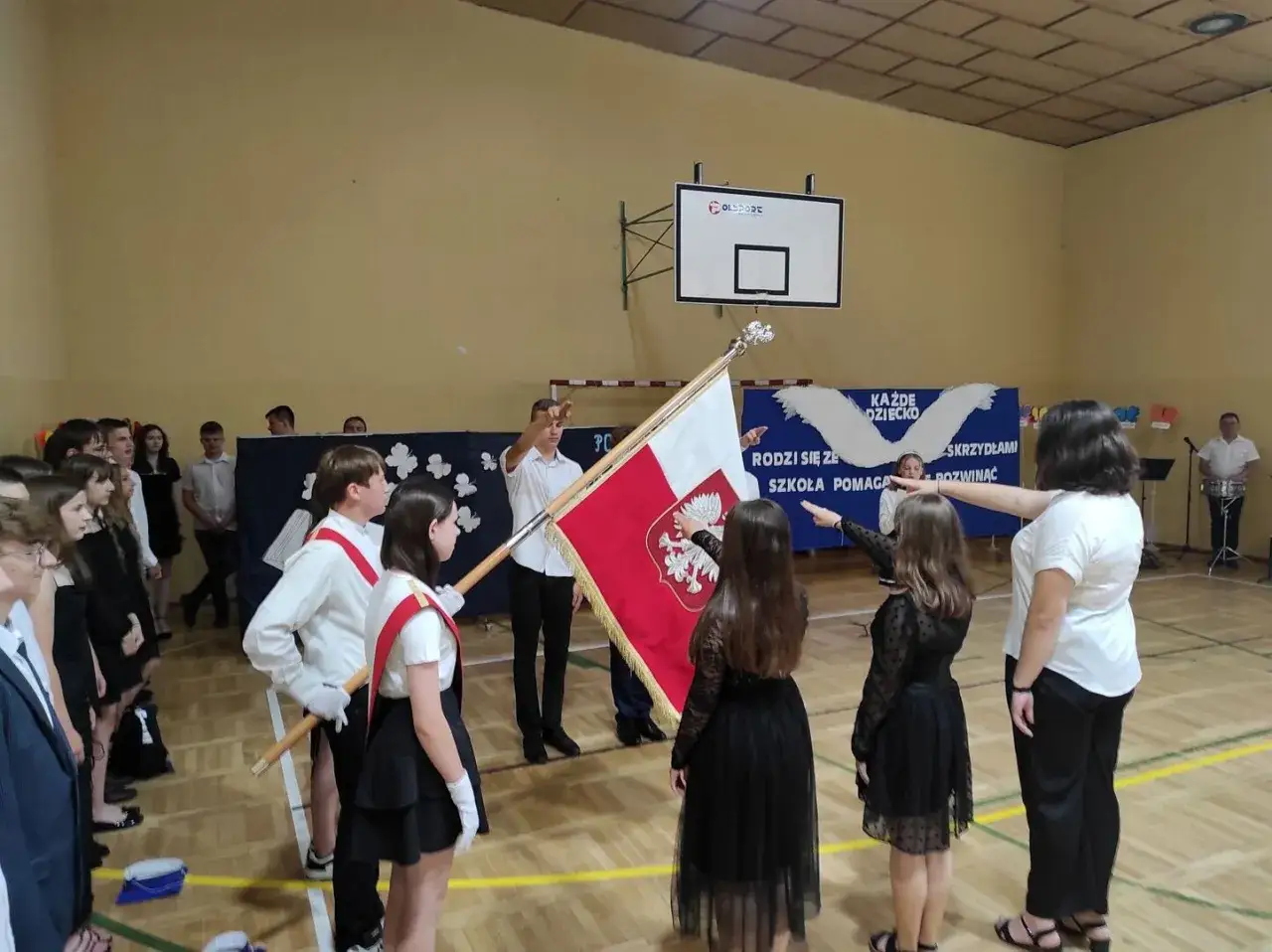 Uroczystość zakończenia roku szkolnego. Uczniowie w galowych strojach salutują sztandarowi.