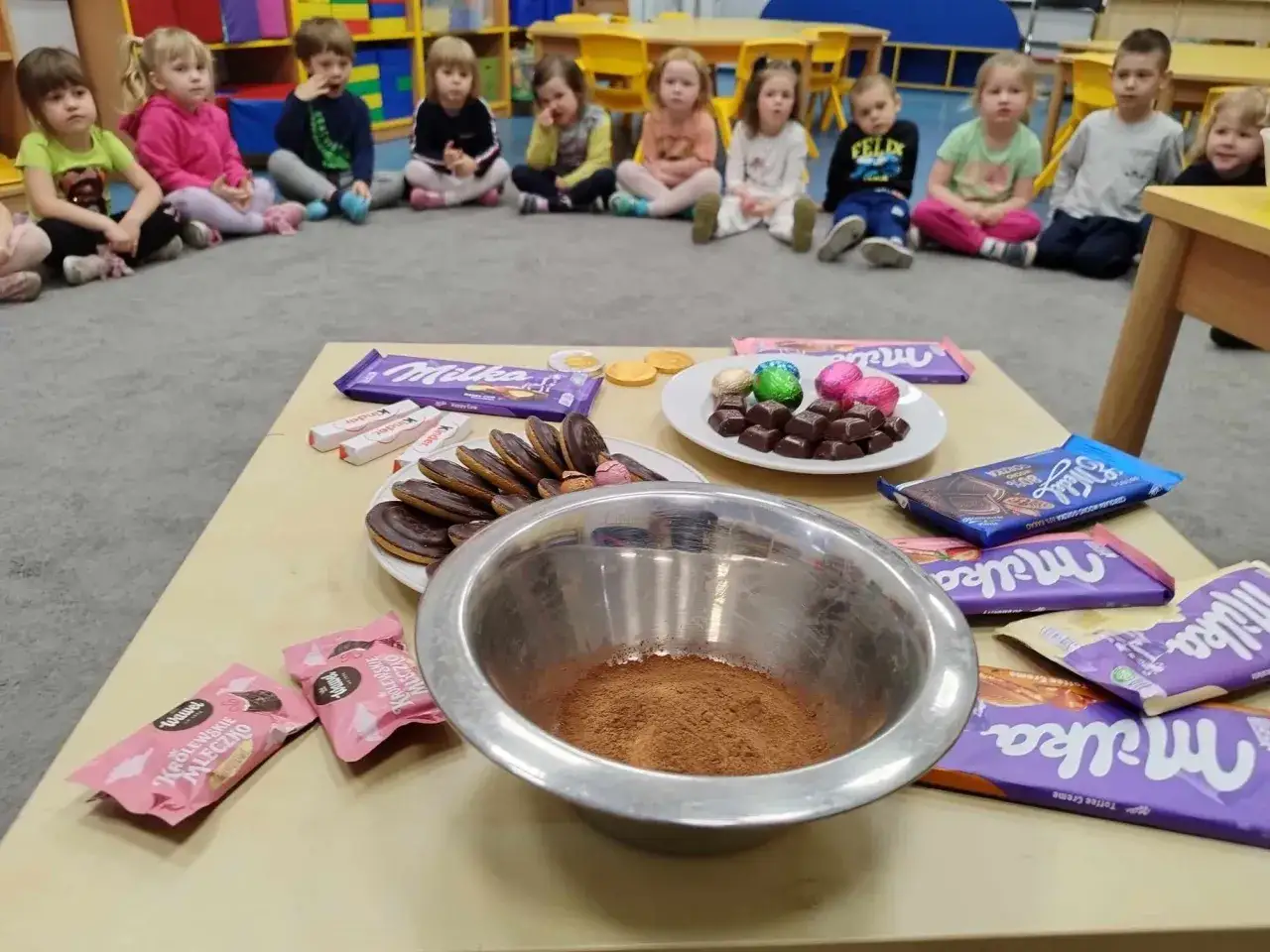 Dzieci w przedszkolu czekają na dzień czekolady. Na stole czekają słodkości: Milka, Kinder, ciastka i kakao.