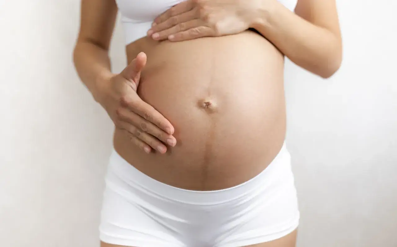 Brzuch kobiety w ciąży z widoczną ciemną linią linea negra biegnącą od pępka w dół. Dłonie delikatnie dotykają brzucha.
